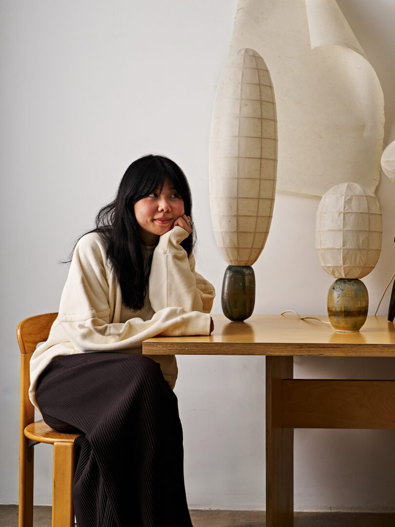 A woman with long black hair wearing a light-colored sweater and dark skirt sits at a wooden table, resting their chin on their hand, next to two tall decorative lamps.