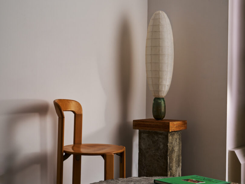 A wooden chair sits next to a pedestal holding a tall, oval-shaped lamp in a minimalistic room. A green book is partially visible on the table in the foreground