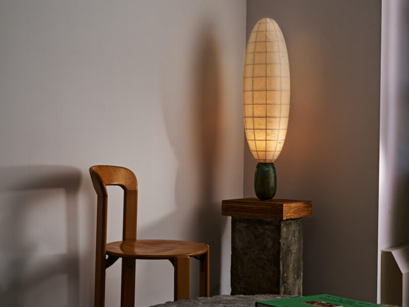A wooden chair sits next to a pedestal holding a tall, oval-shaped lamp lit in a minimalistic room. A green book is partially visible on the table in the foreground
