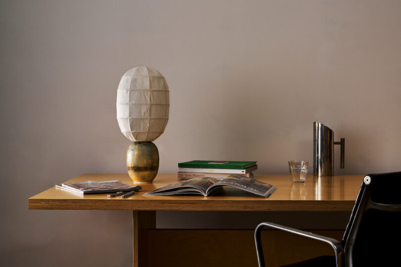 A minimalist workspace featuring a wooden desk with an open book, a stack of closed books, a small lamp with a spherical shade, a glass cup, and a modern black chair