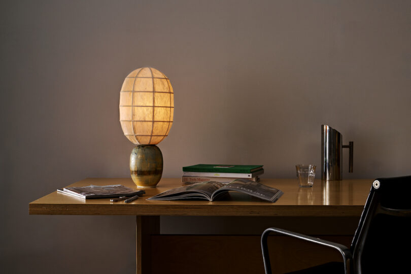 A minimalist workspace featuring a wooden desk with an open book, a stack of closed books, a small lamp with a spherical shade turned on, a glass cup, and a modern black chair