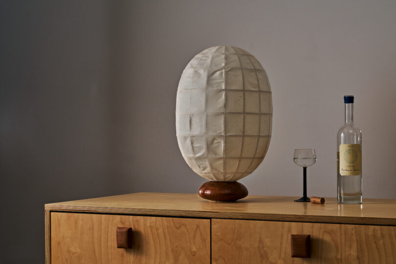A white, oval-shaped lamp on a wooden stand is placed on a wooden dresser. To the right, there's a bottle of clear liquid, a small glass, and a cork. The background is a plain wall