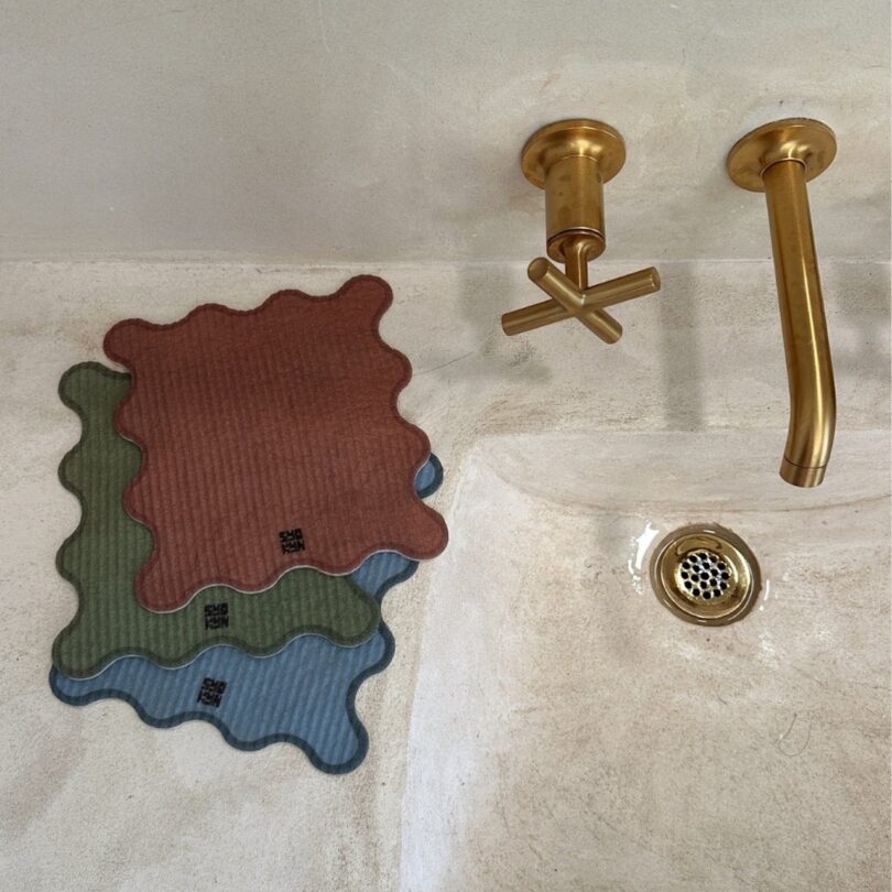 Three wavy-edged reusable paper towels —one brown, one green, and one blue—are placed near a gold-colored faucet and drain on a beige surface