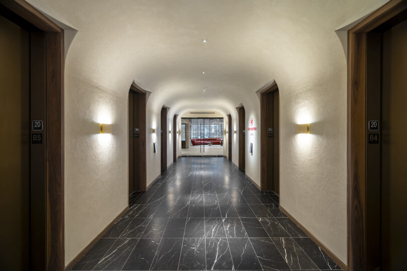 A hallway with arched ceilings, stone floors, and doors labeled "B5" and "B4" on either side, leading to a glass door at the end with a red sofa visible through the glass.