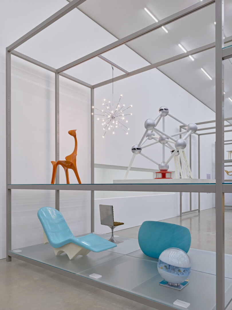 Display of various modern design objects set on metal shelving, including a blue chair, orange giraffe figure, small model of an atom-like structure, and a spherical lamp