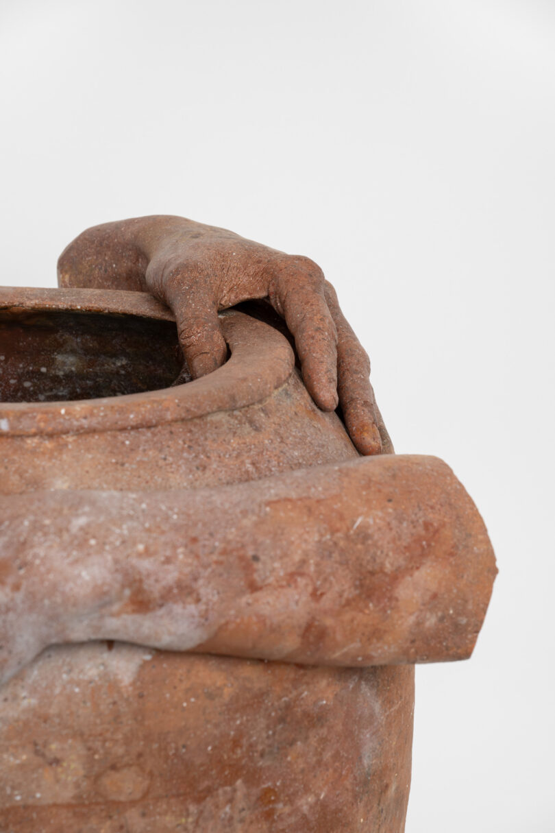 Sculpture of hands rests on opening of clay vessel