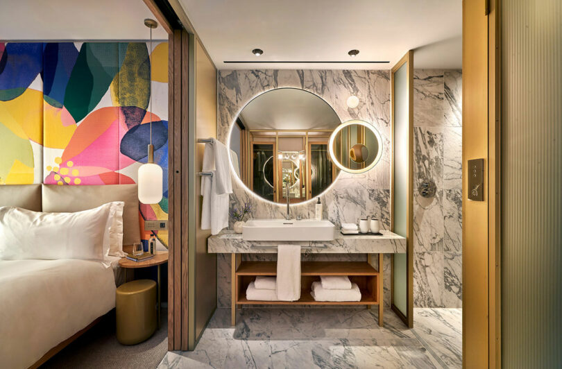 Modern BoTree Hotel room bathroom featuring a marble sink counter with round mirrors, colorful wall art, and a neatly made bed partially visible. The space is illuminated with warm, soft lighting.