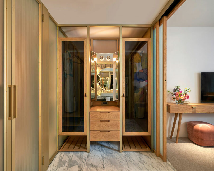 A modern dressing area at BoTree Hotel features wooden framed mirrors, a marble floor, and a vanity table with drawers. A bouquet of flowers adorns a side table next to the TV.