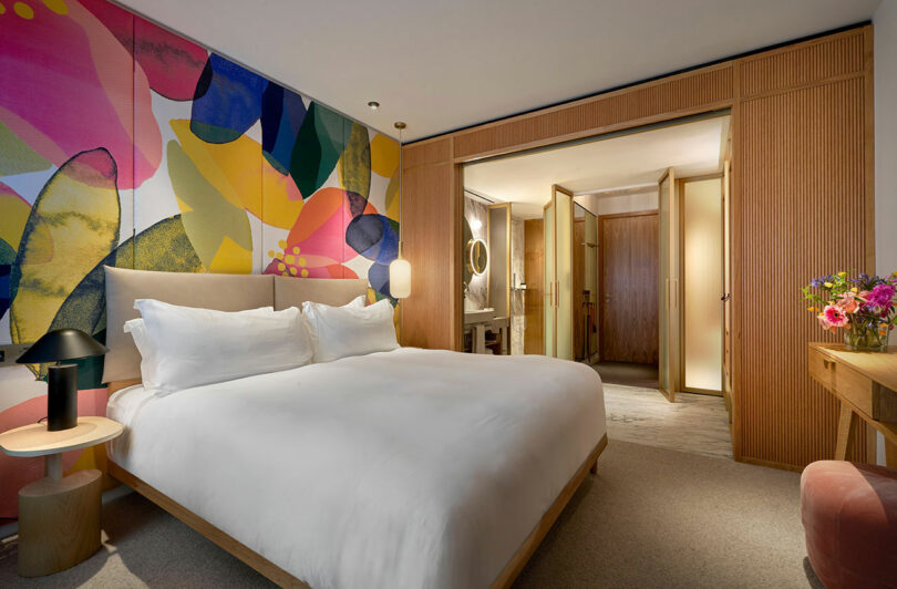A modern BoTree Hotel room with a large bed, colorful floral mural on the wall, bedside tables, flowers on a desk, and an open view into a stylish bathroom.