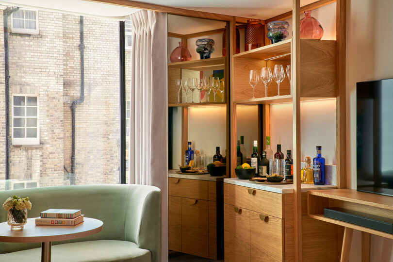 A modern home bar setup with wooden cabinets, glass shelves displaying bottles and glasses, a green armchair, and a small table with books from the BoTree Hotel. A window offers a view of a brick wall outside.
