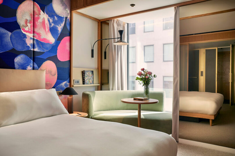 A modern BoTree Hotel room featuring a double bed, a green couch, a small table with a vase of flowers, and a floor-to-ceiling window with an enchanting city view outside.
