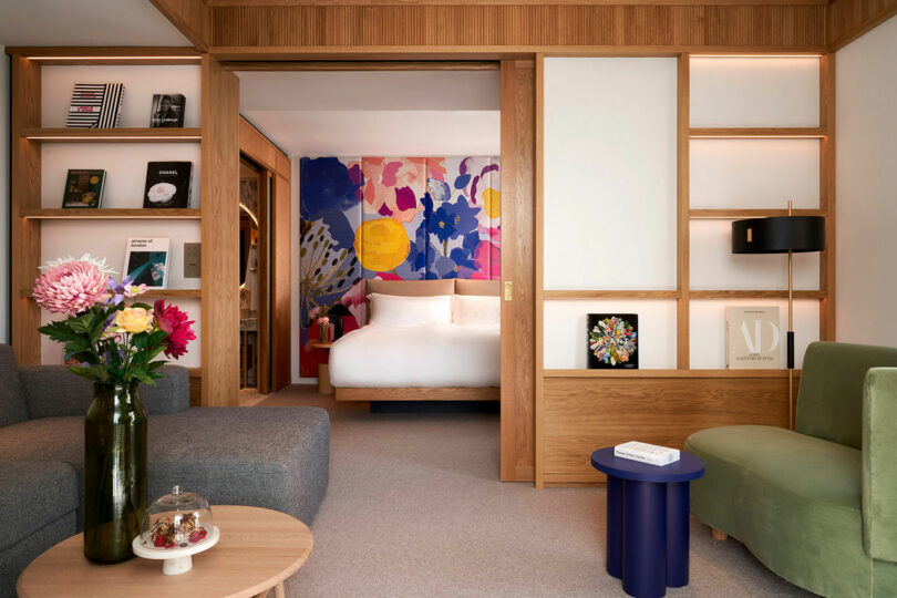A modern hotel room at the BoTree Hotel features a bed, colorful floral mural, gray sectional sofa, green armchair, and bookshelves. A vase of flowers and a cake stand adorn the coffee table in the foreground.