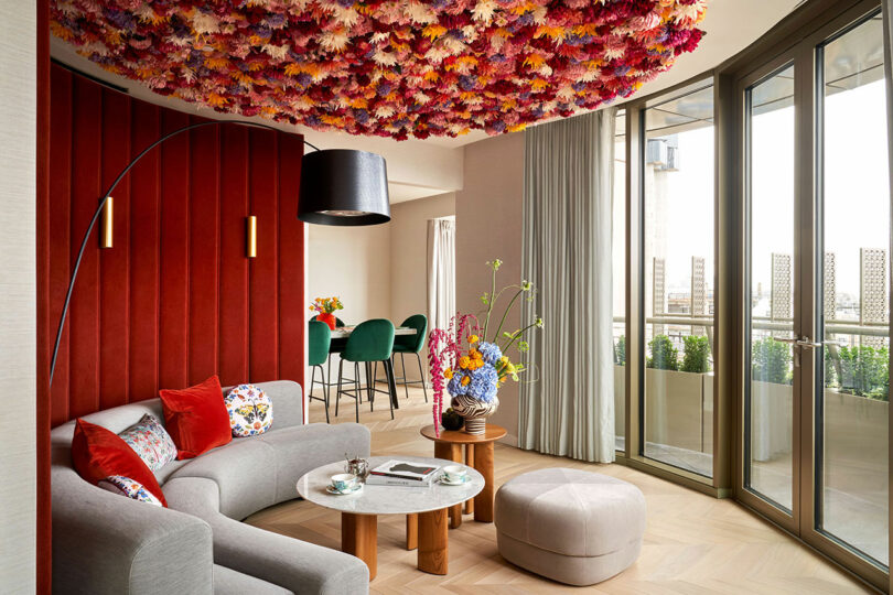 A modern living room at the BoTree Hotel features a floral ceiling, a red accent wall, a grey sofa with colorful cushions, a round marble coffee table, and large windows. The dining area has green chairs and a table by the window, perfect for enjoying the view.