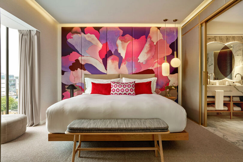 A modern BoTree Hotel room with a king-sized bed, vibrant floral mural, red and white pillows, a bench at the foot of the bed, and a bathroom with marble accents visible through a sliding door.