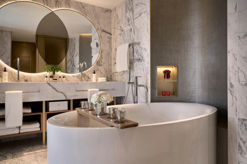 A modern bathroom at BoTree Hotel features a marble motif, a freestanding oval bathtub, a wood bath tray with flowers and toiletries, large round mirrors above the double sinks, and lit shelves adorned with decor.