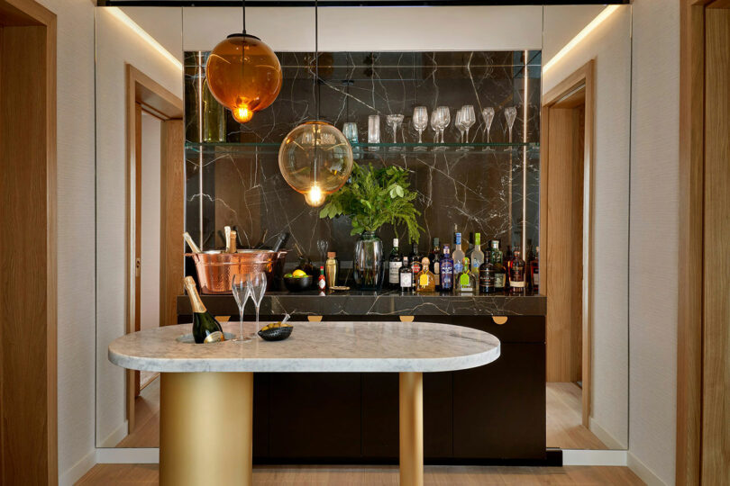 A modern home bar at BoTree Hotel features a marble countertop, various bottles of alcohol, glasses, a wine bottle in an ice bucket, and hanging amber-colored pendant lights.