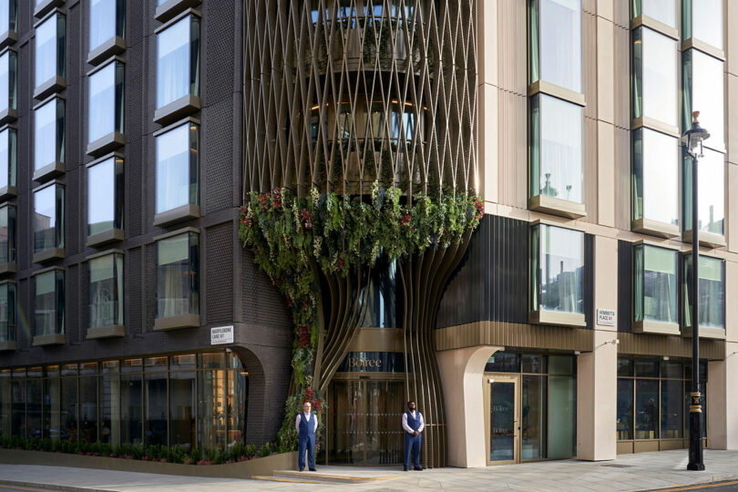 A modern building with a unique architectural design featuring vertical, intertwining metal elements and greenery. Two individuals stand near the entrance of "The BoTree Hotel.
