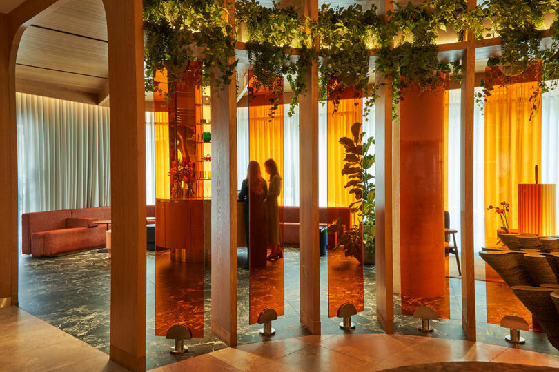 Two people standing near a tall structure with orange illuminated panels and hanging greenery in the modern, stylish indoor setting of the BoTree Hotel, surrounded by assorted seating areas.