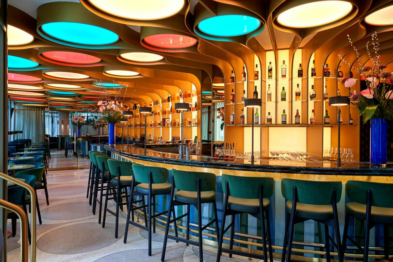 A modern bar at the BoTree Hotel features a curved counter, colorful ceiling lights, and shelves stocked with various bottles. Bar stools with teal seats line the counter. Decorative flowers are placed at either end of the bar.