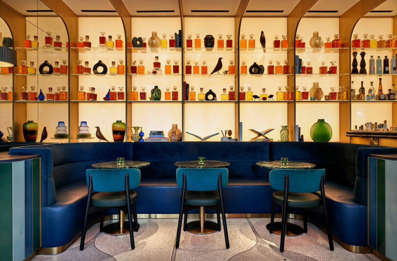 A stylish bar at the BoTree Hotel features a curved blue seating area, three small round tables with chairs, and a back wall adorned with shelves filled with various bottles and decorative items.