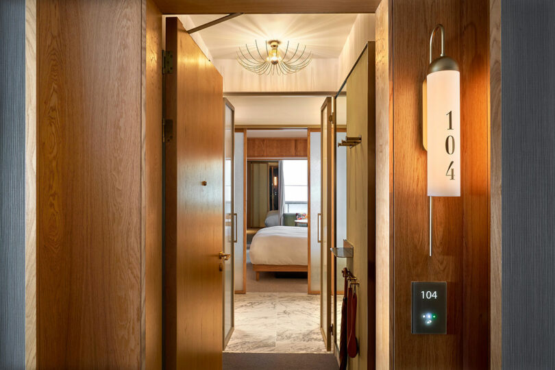 A brightly lit entrance at BoTree Hotel features wooden doors and walls. Room number 104 is prominently displayed beside the door. Inside, the room is visible with a neatly made bed and a chandelier adding an elegant touch.