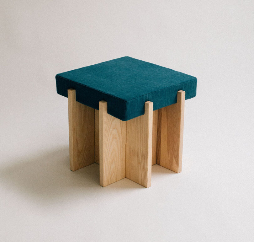 A wooden stool with a blue cushioned seat. The stool has a cross-shaped base and a minimalist design, set against a plain background.