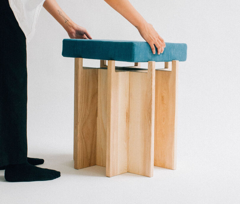 A person places a blue-cushioned stool on top of a wooden structure with a white background.