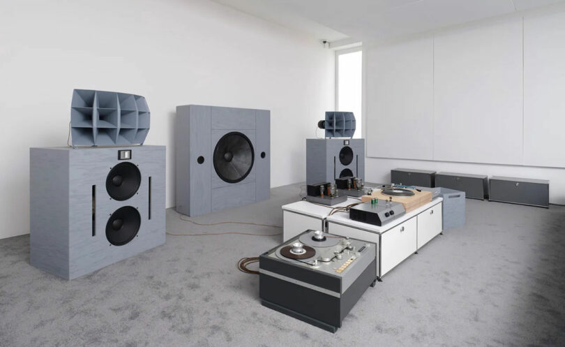 A room features a large audio setup with multiple grey speakers, a turntable, and sleek audio equipment arranged on modern white and grey furniture. The walls are pristine white, complementing the carpeted grey floor.