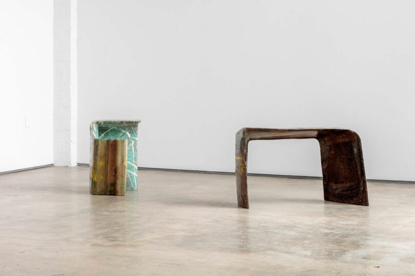 Two modern sculptural furniture pieces in a minimalist setting—one made of green marble and the other of rustic brown material—are placed on a polished concrete floor against a pristine white wall background.