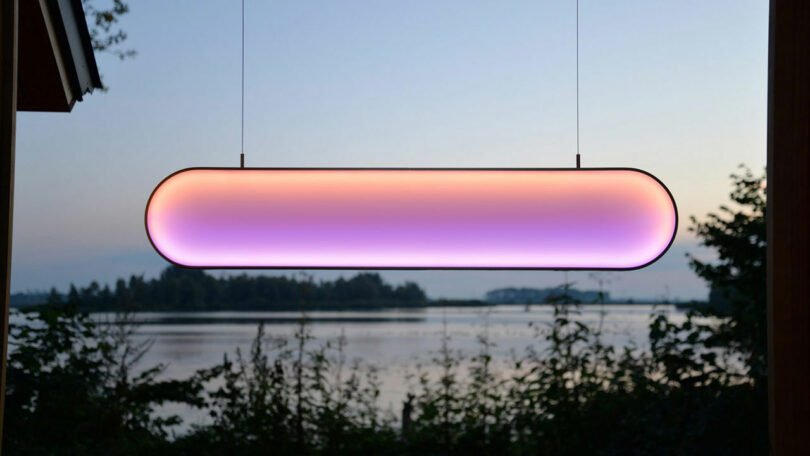 Oblong-shaped, color-changing LED light fixture suspended outdoors at dusk by Benni Allan, with a serene lake and trees in the background.