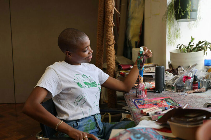 A woman wearing a white t-shirt and blue jeans sits at a cluttered table, holding a paintbrush and looking at their artwork in a studio setting.