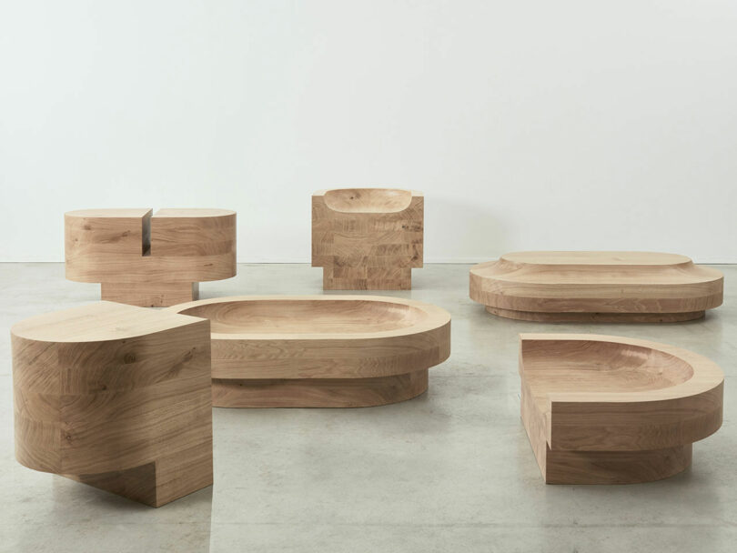 A collection of uniquely shaped, minimalistic wooden furniture pieces by Benni Allan is displayed in a spacious room with a concrete floor and a plain white background.