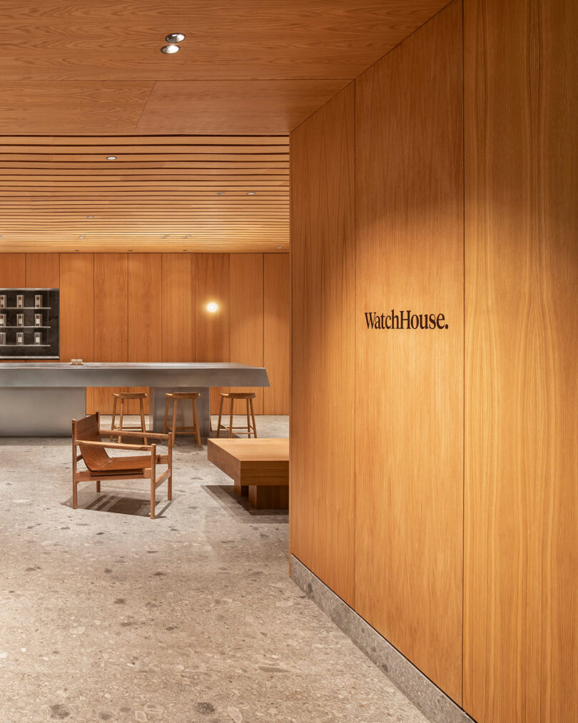 A modern interior designed by Benni Allan features light wooden walls and a stone floor. A sign reading "WatchHouse" is mounted on the right wall, and the space includes minimalist furniture and a counter in the background.