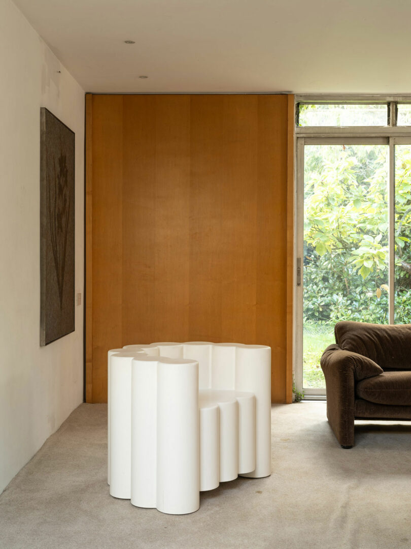 A modern living room features a unique white sculptural table, a brown couch, a wooden accent wall, a large window, and wall-mounted artwork. Greenery from the outside is visible through the window.