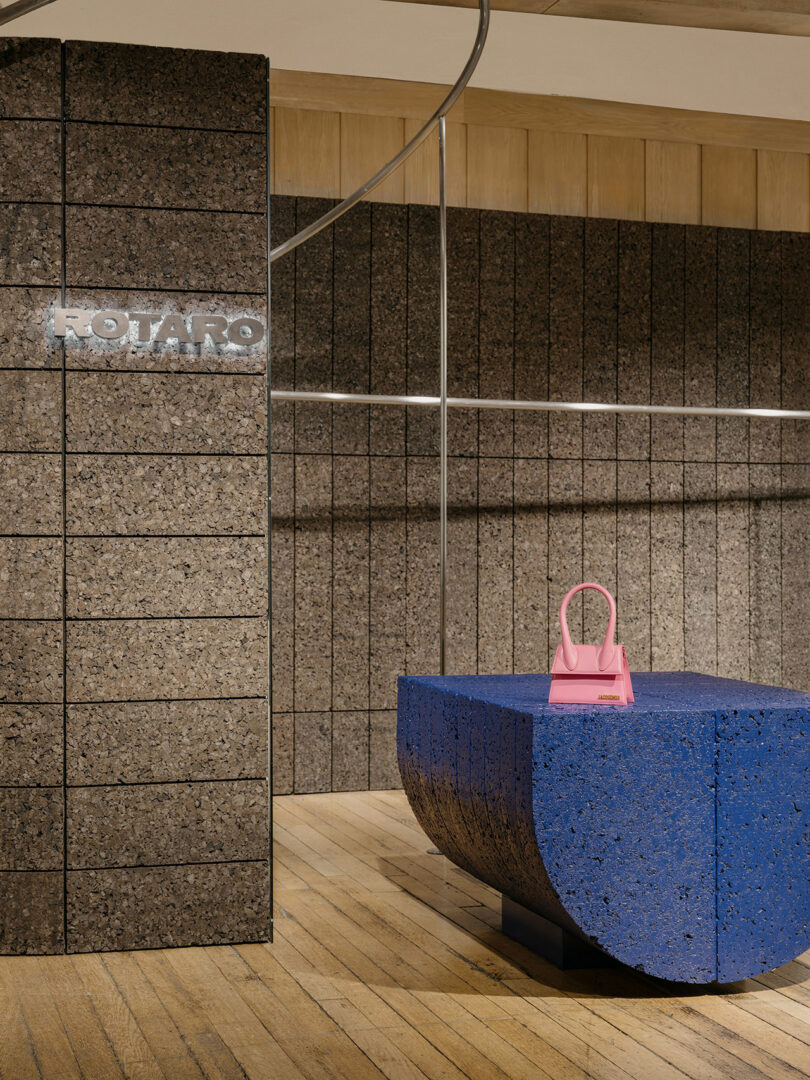 A pink handbag is displayed on a textured blue pedestal in a minimalist store featuring wooden flooring and brown tiled walls. A sign with the text "ROTARO" is visible on the wall.