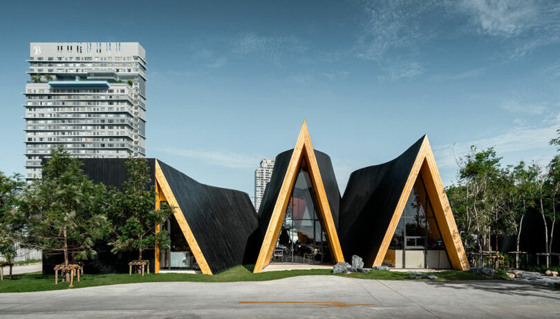 A modern building with sharp, triangular peaks and wooden accents stands before a blue sky with a tall apartment complex in the background.