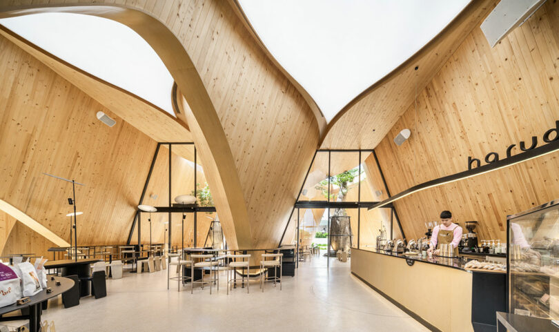 Interior view of a modern café with tall, curved wooden walls, ample seating, large windows, and a barista preparing drinks behind the counter.