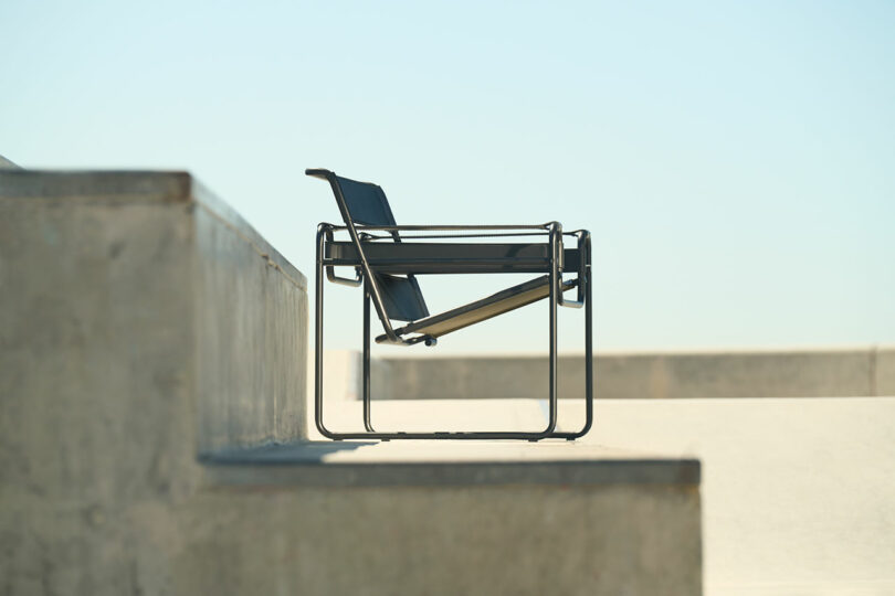 A modern black metal chair placed on a concrete surface with clear blue sky in the background. Steps and a concrete wall are visible to the left.