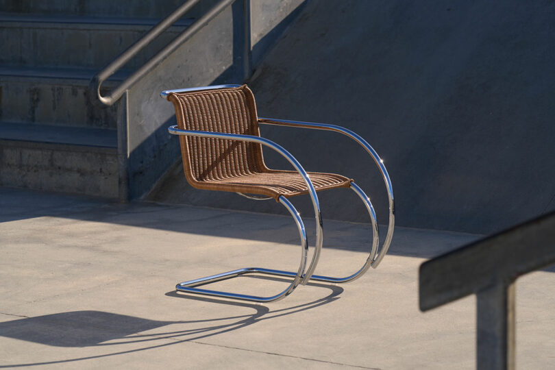 A single, modern chair with a brown woven seat and metal frame sits on a concrete surface near stairs with metal railings.