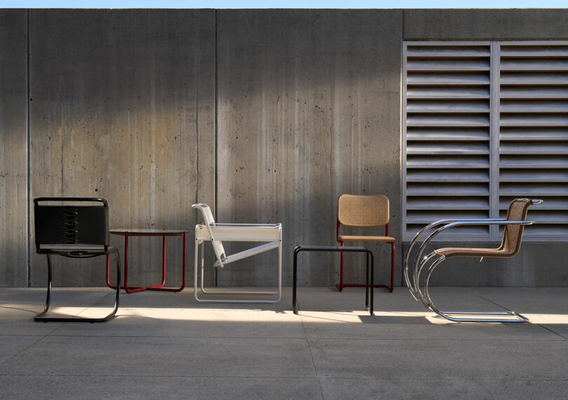 A row of five modern, minimalist chairs against a concrete wall with a ventilation panel. The chairs are made of various materials including metal, wood, and plastic, each with a unique design.