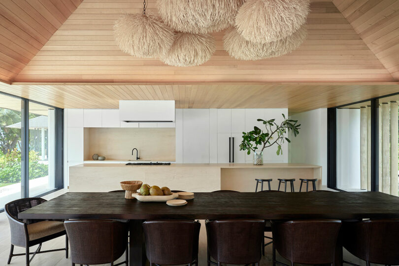 Modern kitchen and dining area with wooden ceiling and large hanging light fixtures. A long dark wooden dining table with eight chairs is in the foreground, and the kitchen features white cabinetry.