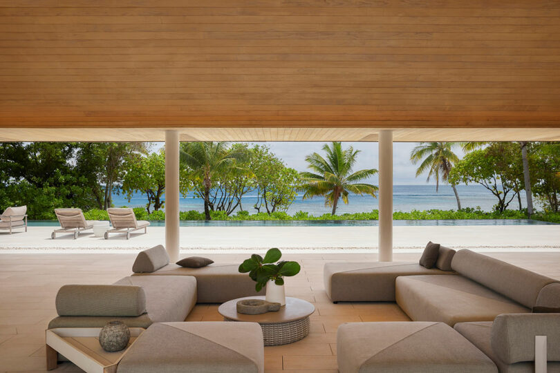 Open-air lounge with modern furniture and a low table, under a wooden ceiling. View of outdoor loungers, lush greenery, and the ocean in the background.