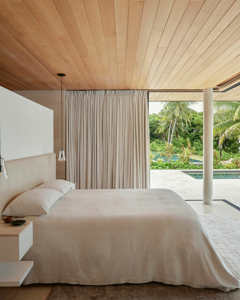 A modern bedroom with a wooden ceiling, white bedding, a curtain, and a view of a pool and tropical vegetation through sliding glass doors.