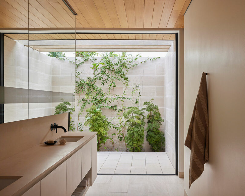 Modern bathroom with a minimalist design featuring a countertop sink, black faucet, towel rack with a hanging checkered towel, and a large glass window looking out to a small outdoor green space.