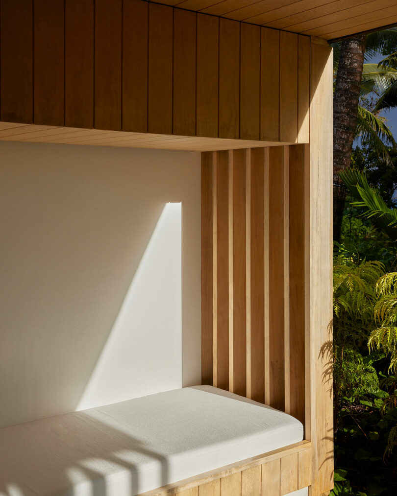 A wooden alcove featuring a cushioned bench seat. Sunlight streams through a gap, creating a bright line on the white wall. Lush greenery is visible outside.