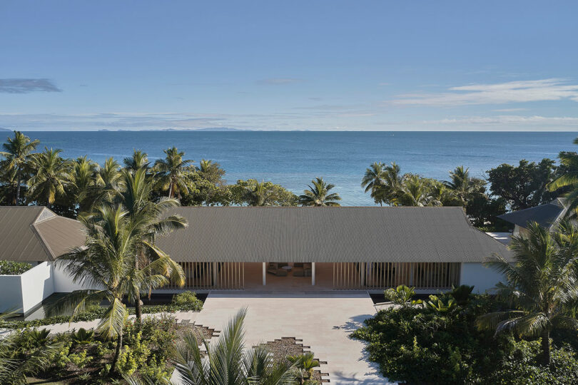 A modern building with a pitched roof is surrounded by tropical palm trees, located near a calm, blue ocean under a clear sky.