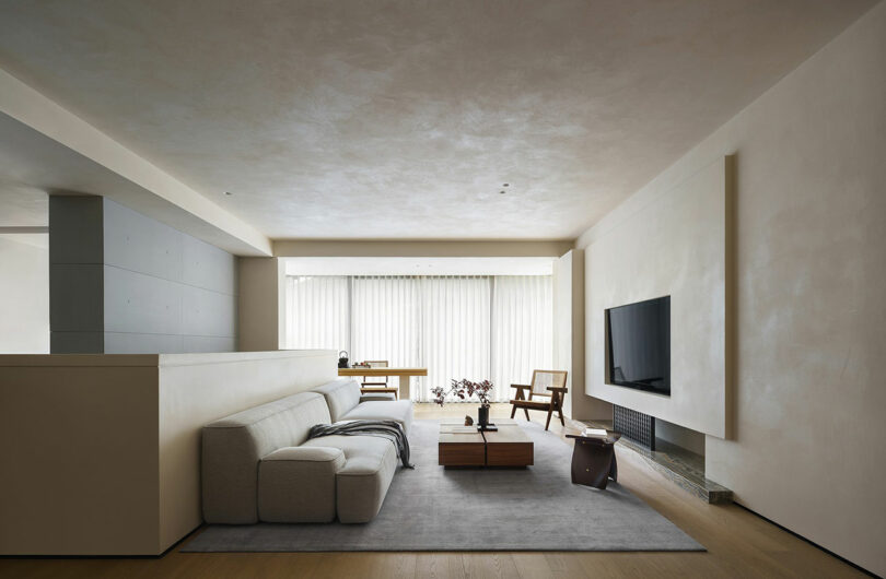 A minimalist living room with a light gray sectional sofa, a wooden coffee table, a wall-mounted TV, and a large window with sheer curtains allowing natural light to fill the space.