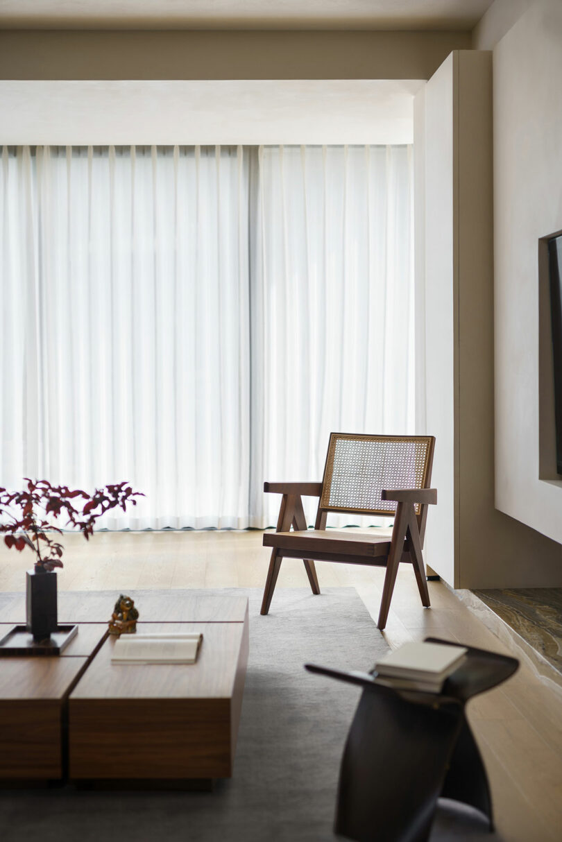 Modern living room with a wooden armchair next to a large window with sheer curtains. A coffee table with minimal decorations and a book stand are also present.