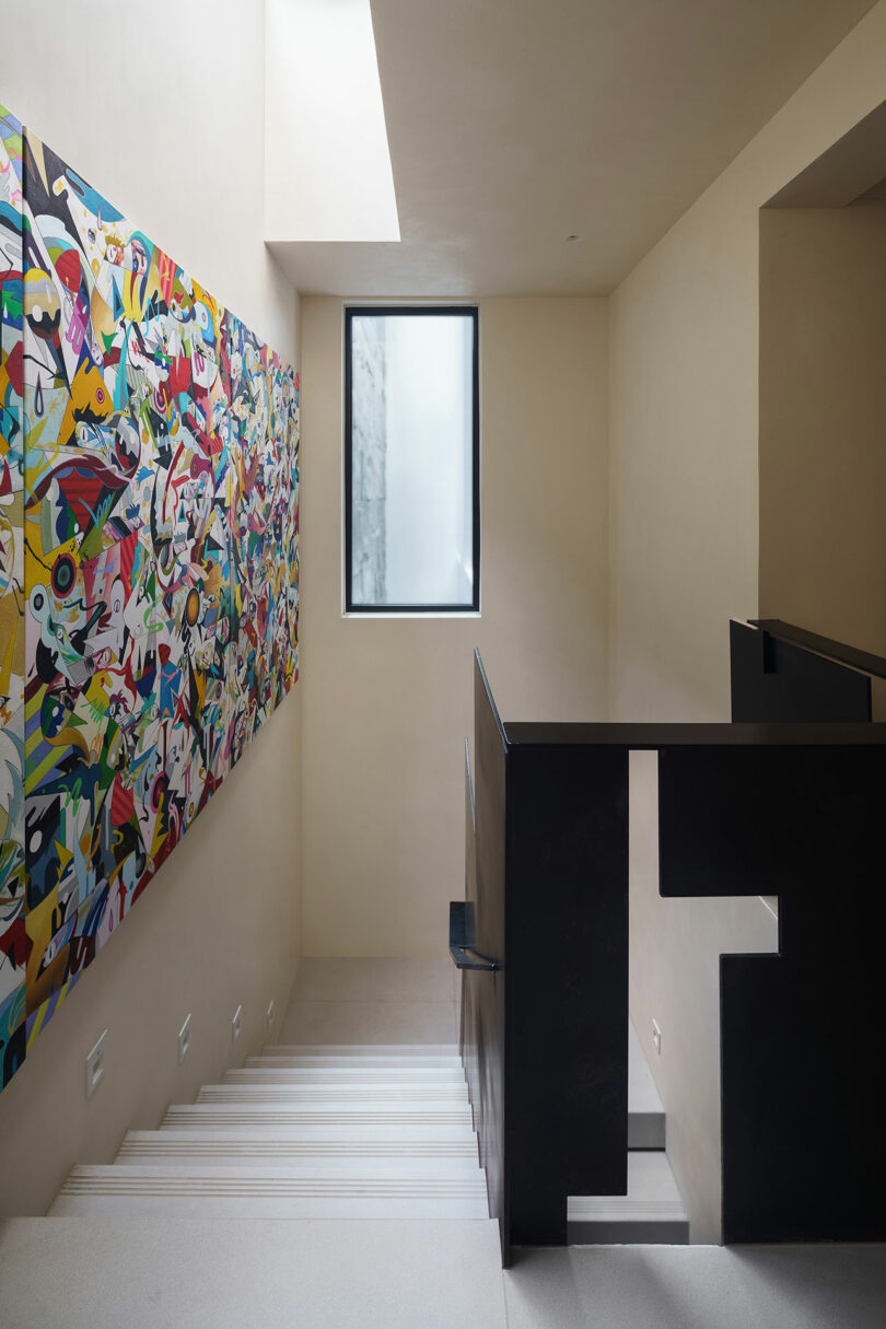 A staircase with white steps and beige walls is adorned with a colorful abstract painting and illuminated by a rectangular window. The stair railing is black with a modern design.