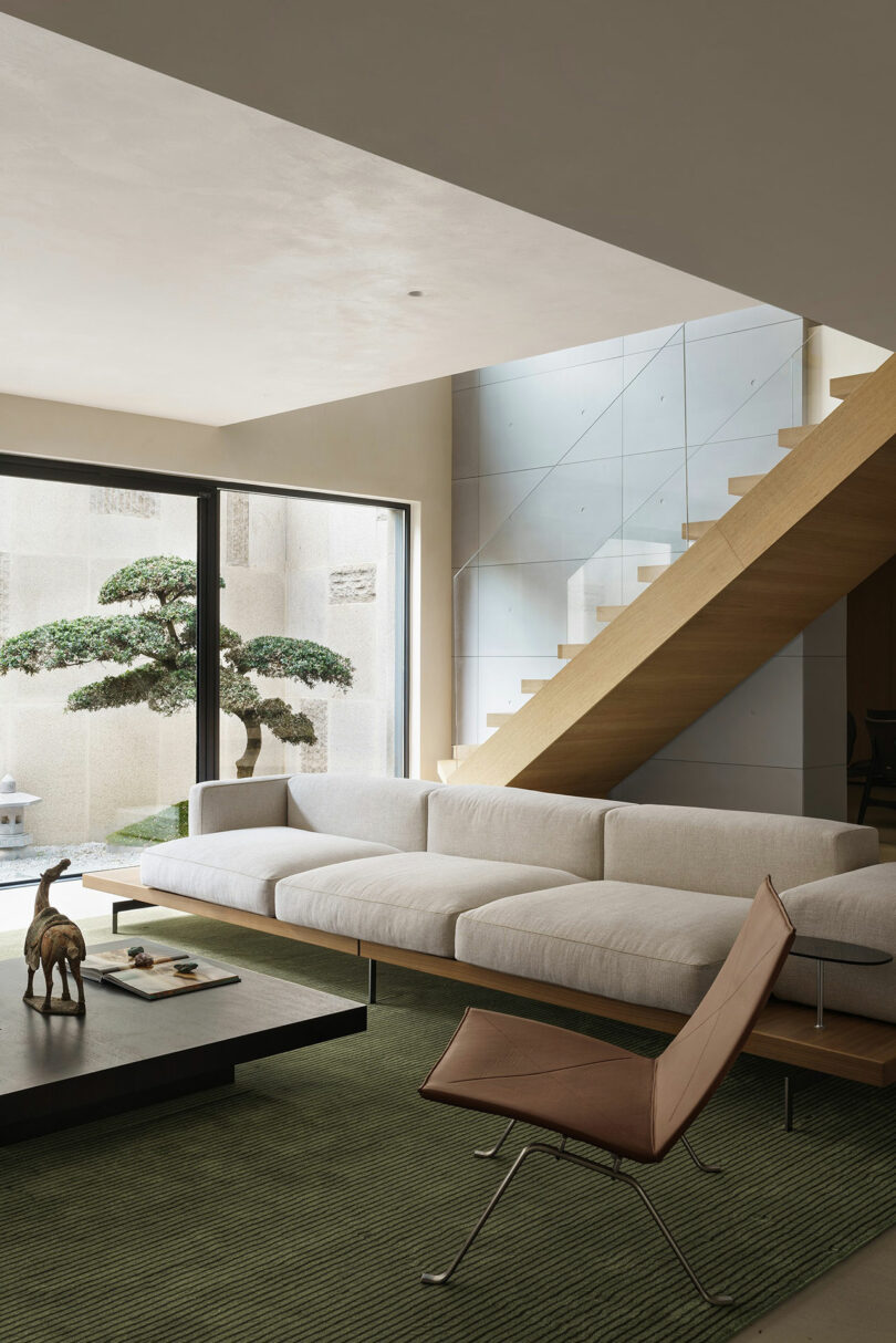 Modern living room with a white sofa, brown chair, and coffee table with decor items. Large windows reveal a tree outside. Wooden staircase ascends to the right. Green rug covers the floor.